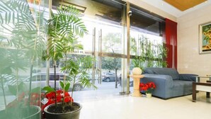 A hotel lobby with a large glass window, potted plants, a grey sofa, and a small table.