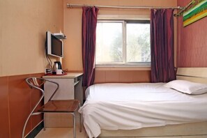 Ein Einzelbett mit weißer Matratze und Holz-Kopfteil, ein kleiner Schreibtisch mit Stuhl, ein an der Wand befestigtes Fernsehgerät und ein Fenster mit Vorhängen.