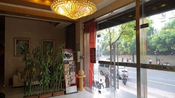 A hotel lobby with a large chandelier, indoor plants, a glass door entrance, and a poster on the wall.
