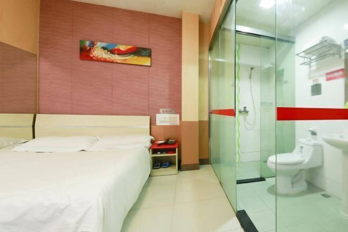 A hotel room with a bed, a small table with a phone and slippers, a glass shower enclosure, and a toilet.