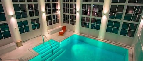 Una piscina interior con vista despejada al área de la piscina, escalones que bajan al agua y una zona de estar con una silla roja.