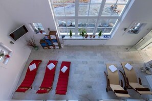 A hotel room with a large window, a TV, a desk, and two red lounge chairs.