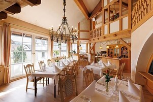 Un comedor con mesas y sillas de madera, una araña y vistas al exterior.