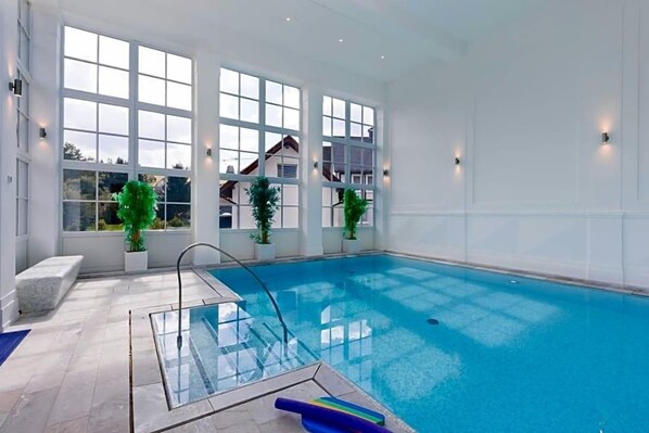 An indoor swimming pool with clear glass walls, potted plants, and a view of a house through large windows.
