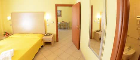 Habitación de hotel con cama, mesita de noche, lámpara y baño con lavabo y espejo.
