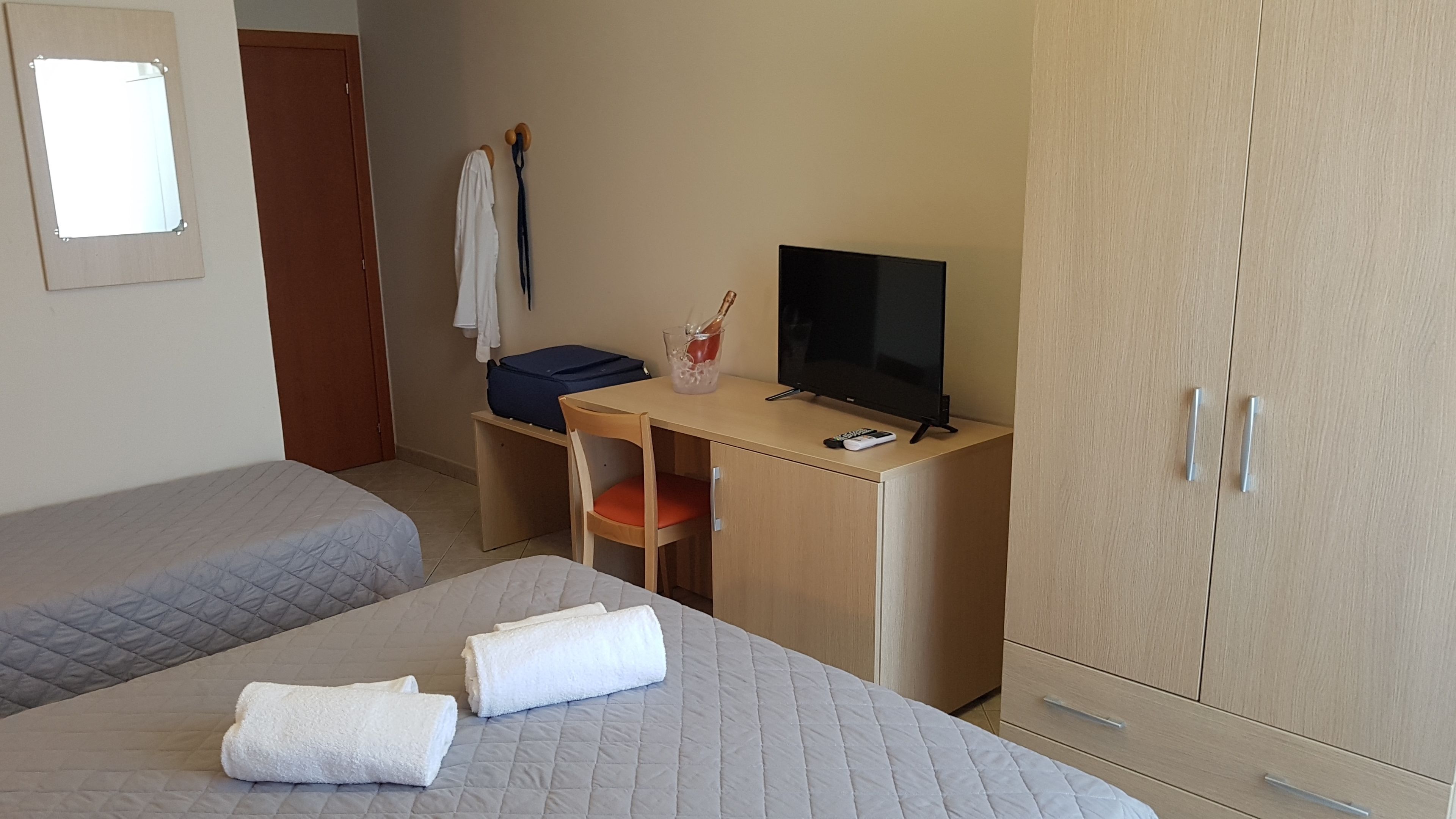 A hotel room with two beds, a desk with a TV, a wardrobe, and a small table with a bottle of champagne.