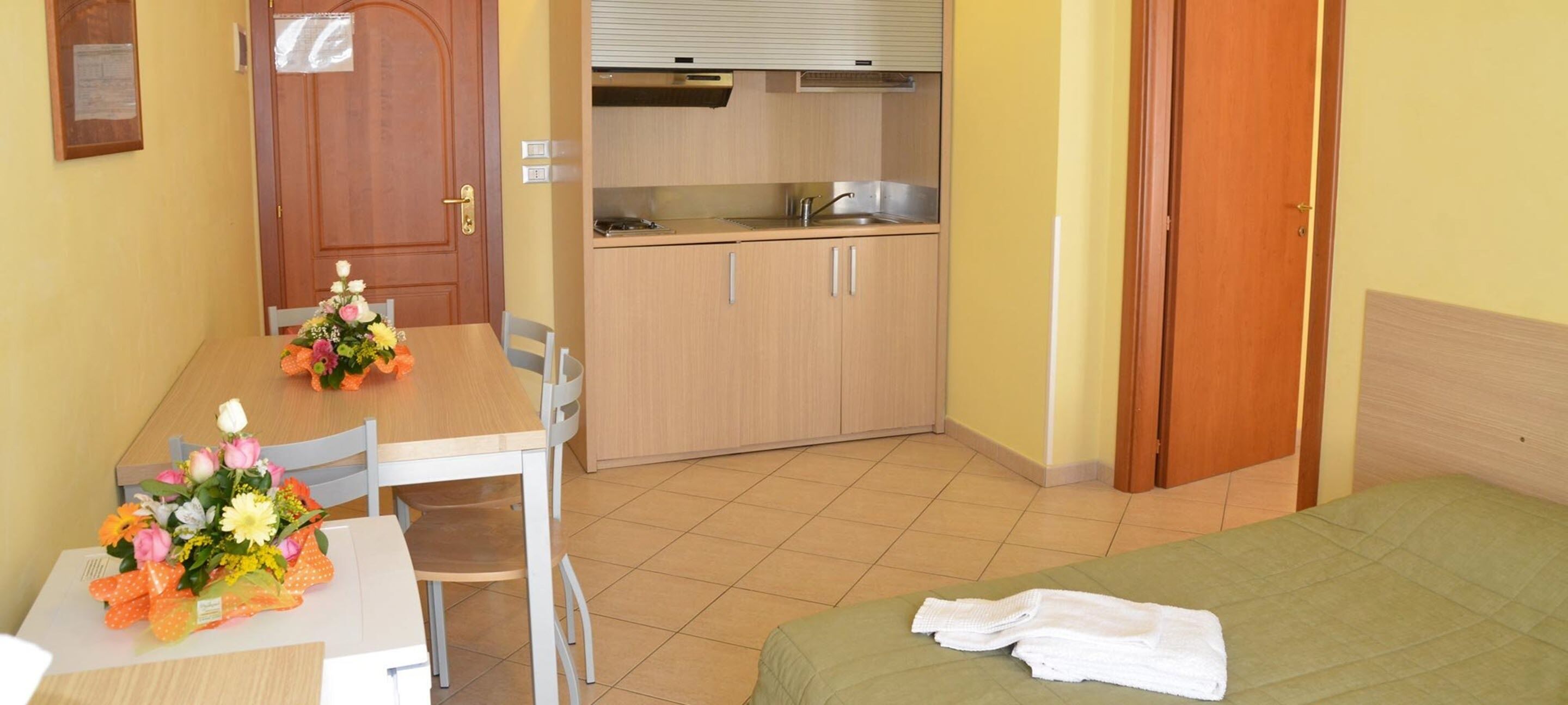 Una camera d'albergo compatta con un letto, una scrivania piccola con una sedia, un angolo cottura e una porta in legno.