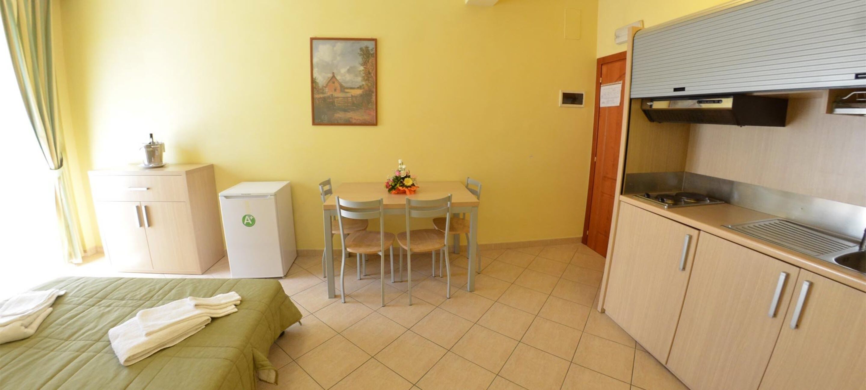 Une chambre d’hôtel compacte comprenant un lit, une petite kitchenette, une table à manger avec des chaises et un tableau au mur.