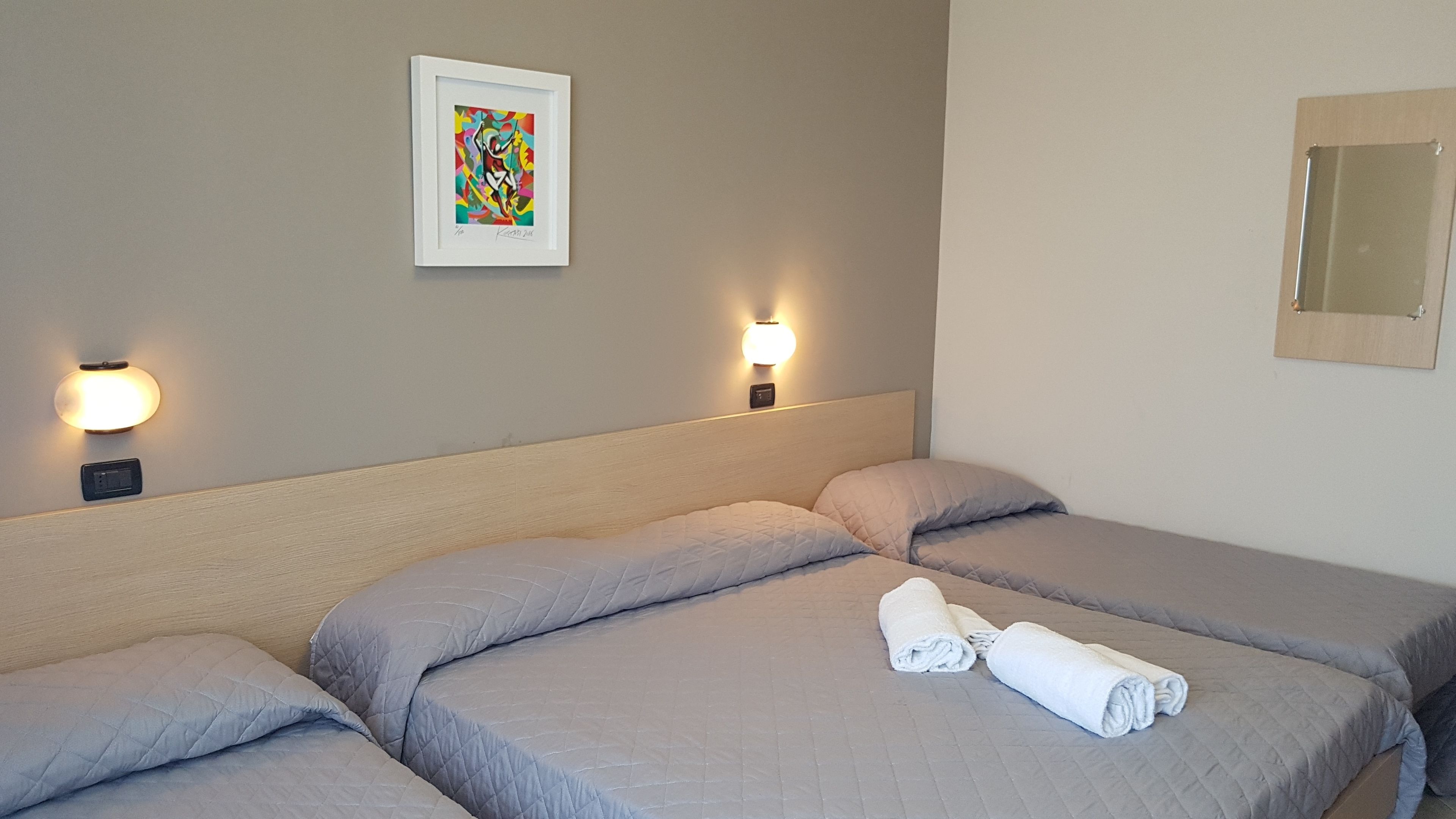 Een hotelkamer met drie bedden, elk voorzien van een quiltdeken en kussens. Aan weerszijden van de bedden zijn wandlampen bevestigd en boven het linkerbed hangt een abstract schilderij in een lijst.