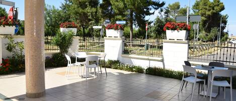 An outdoor patio area with white chairs and tables, surrounded by a tiled floor and a low fence.
