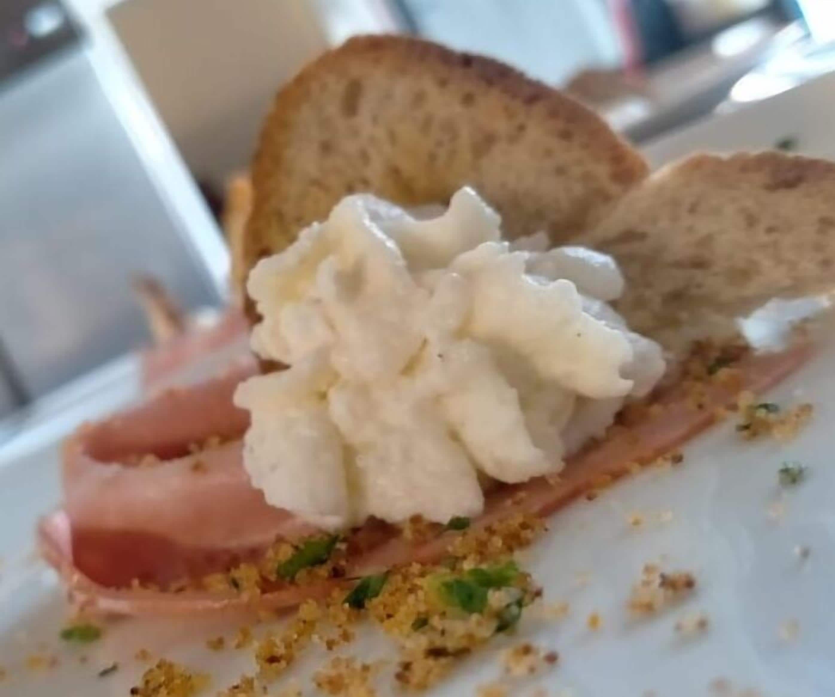 A plate with a slice of ham, a dollop of cream cheese, and crumbled bread.