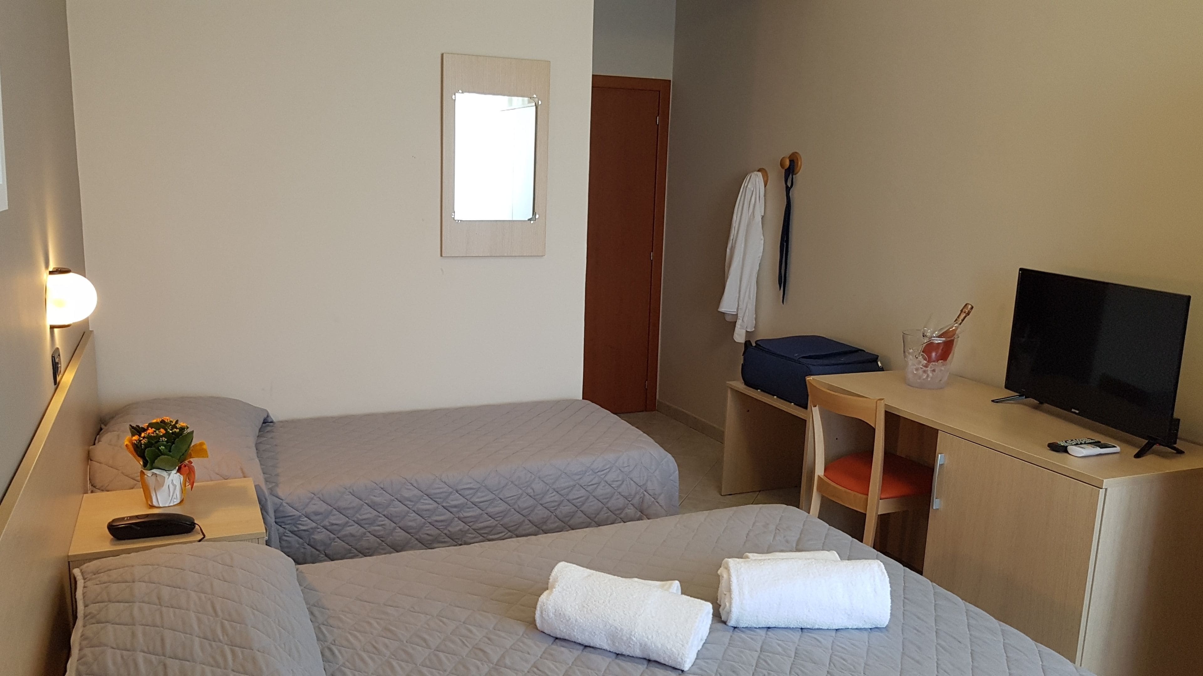 A hotel room with two beds, a desk with a TV, a lamp, a suitcase, and a bouquet of flowers.