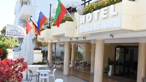 Ein Außenbereich mit Tischen und Stühlen, ein Schild für das Hotel Excelsior und mehrere Flaggen.