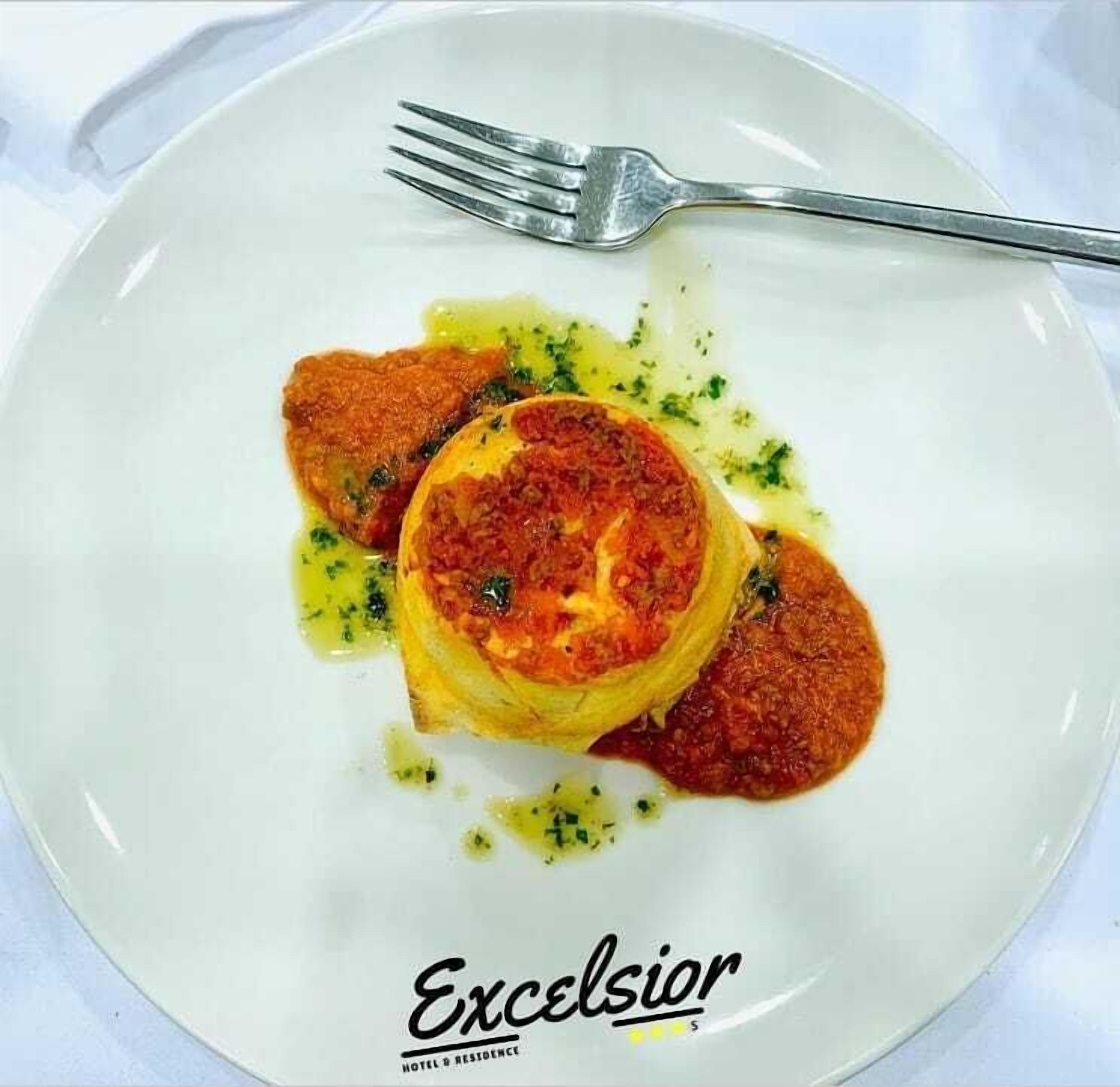 Un plato de comida con un tenedor sobre un plato blanco, con la etiqueta 'Excelsior HOTEL & RESIDENCE'.