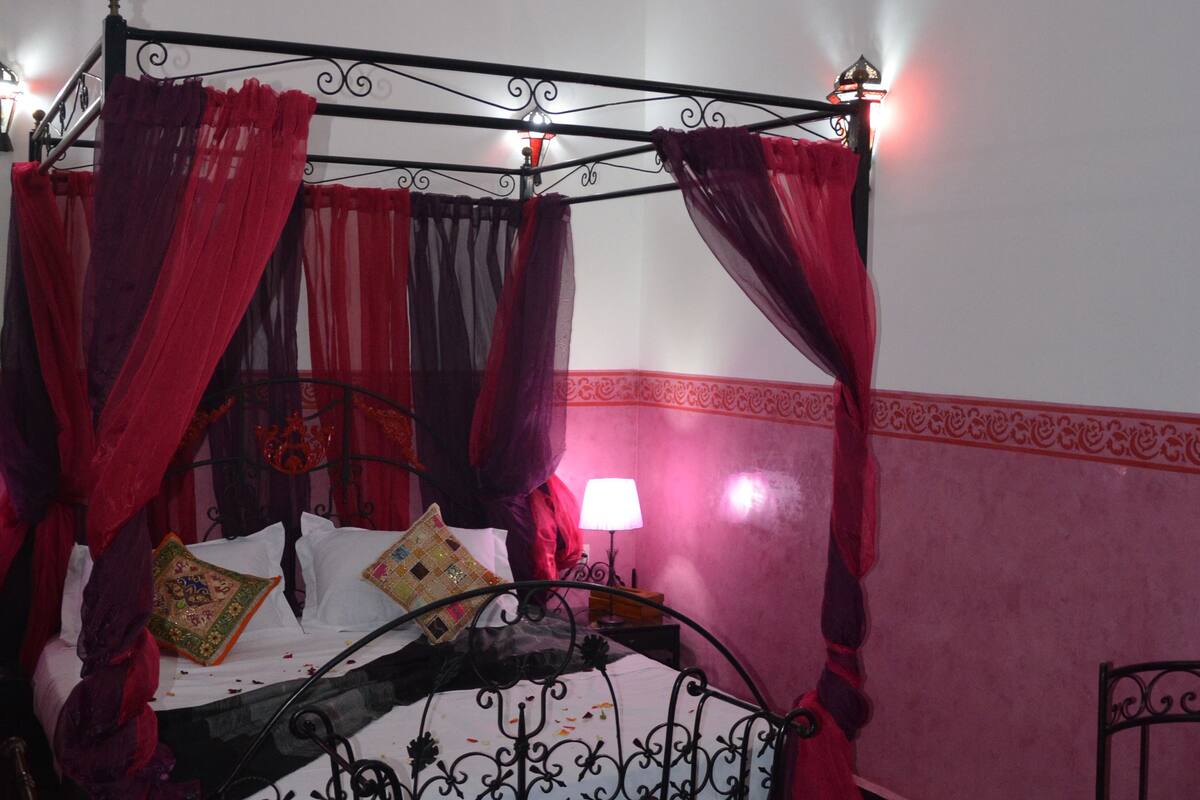 A four-poster bed with red drapes, a small table with a glass, and a chair.