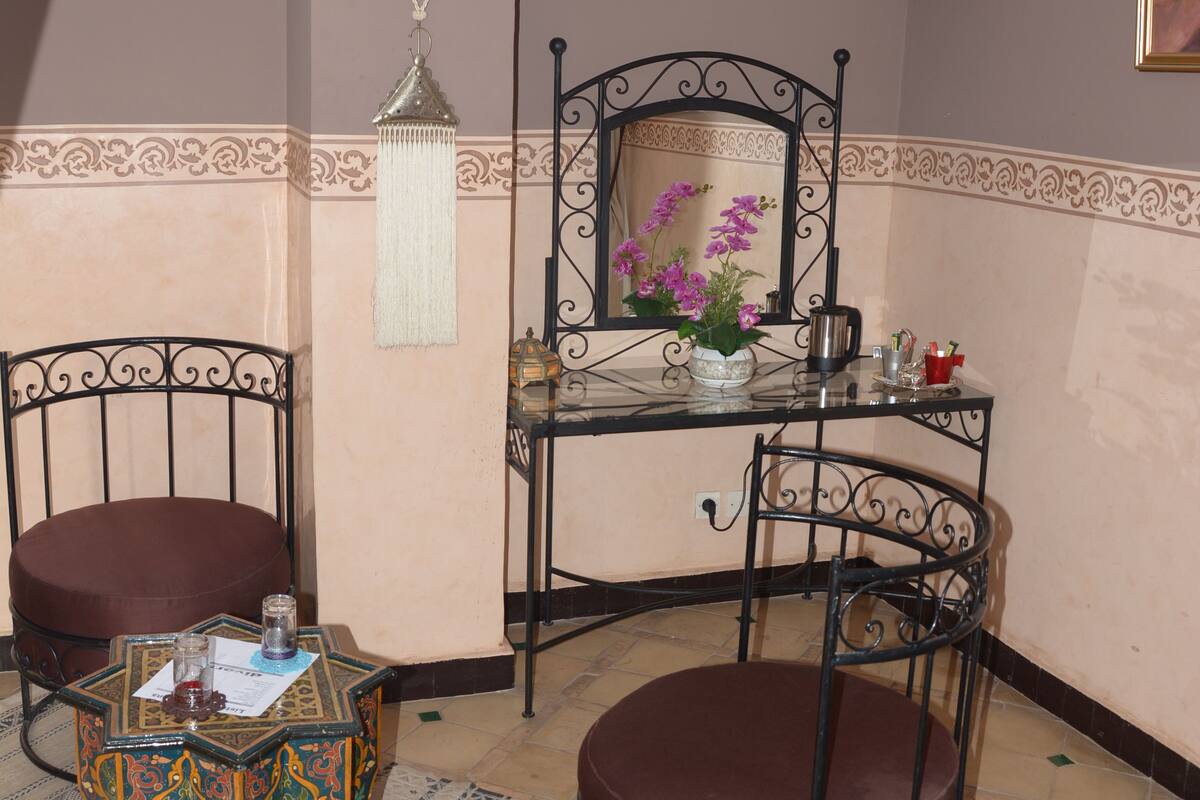 A room with a mirrored vanity, two chairs, a decorative table, and a patterned rug.