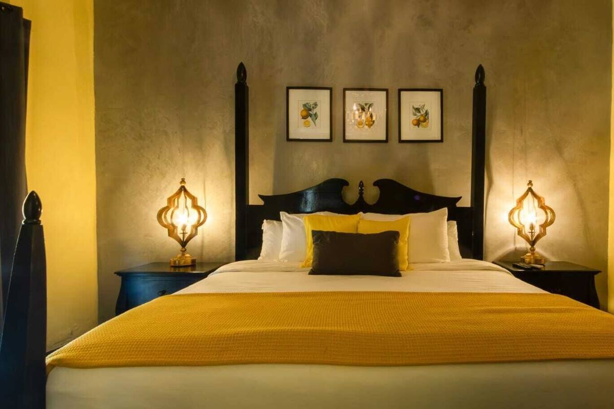 Une chambre d’hôtel avec un grand lit, deux tables de chevet avec des lampes, une tête de lit sombre et trois tableaux encadrés accrochés au mur.