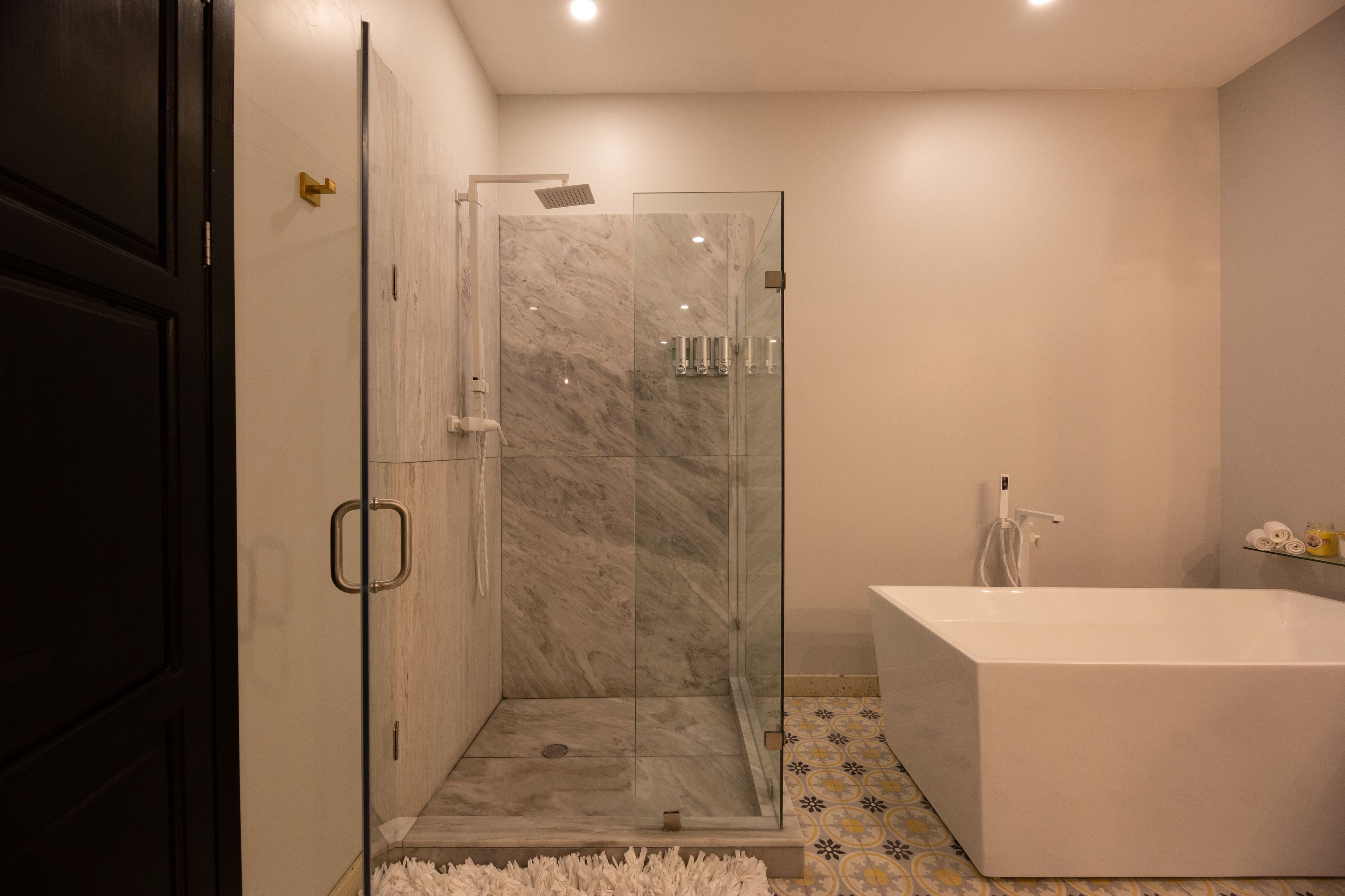 A modern bathroom with a glass-enclosed shower, a white bathtub, and patterned floor tiles.