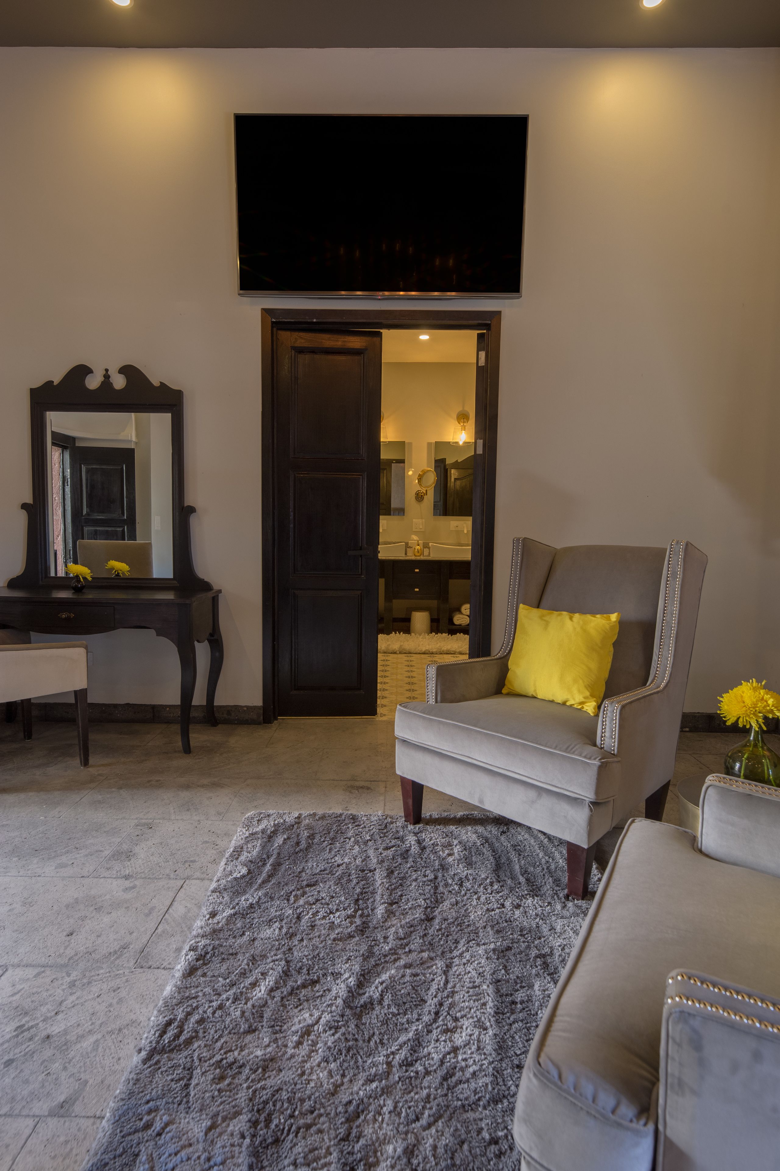 Una sala de estar con un televisor de pantalla plana, una puerta de madera oscura, un sillón gris con un cojín amarillo, un sofá blanco y una alfombra gris.