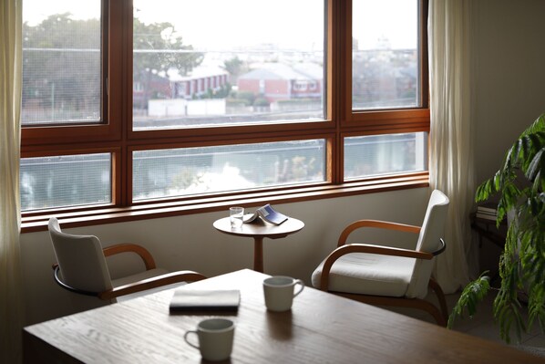 A room with a wooden table, two chairs, a small round table with a glass and a book, and a view of buildings through large windows. - Sango Katase Guest Rooms (Fujisawa)