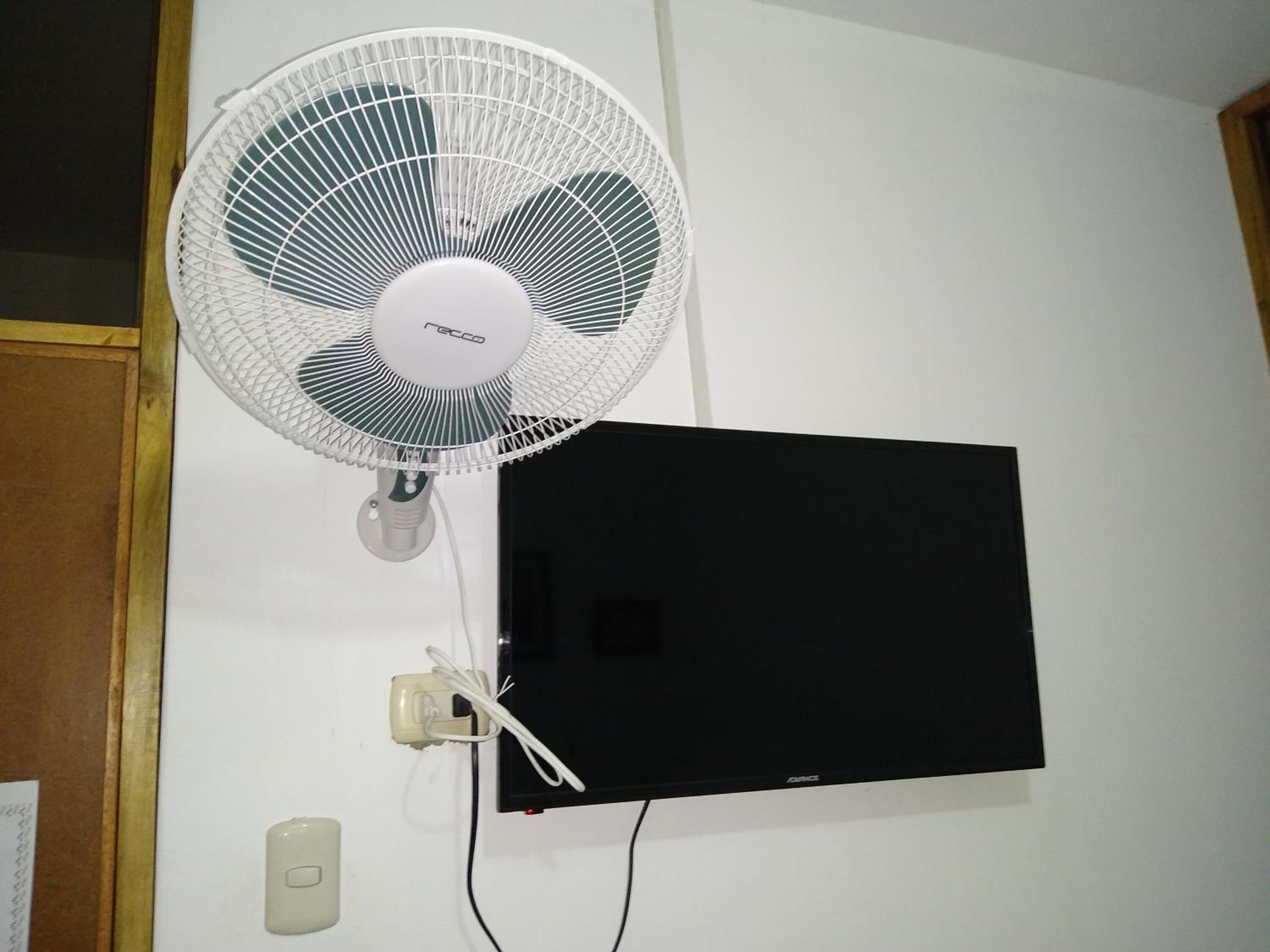 Een zwart, platte flatscreen-televisie aan de muur en een witte ventilator met een snoer.