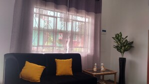 Una sala de estar con un sofá negro, dos cojines amarillos, una pequeña mesita de madera con dos objetos decorativos, y una planta alta en maceta.