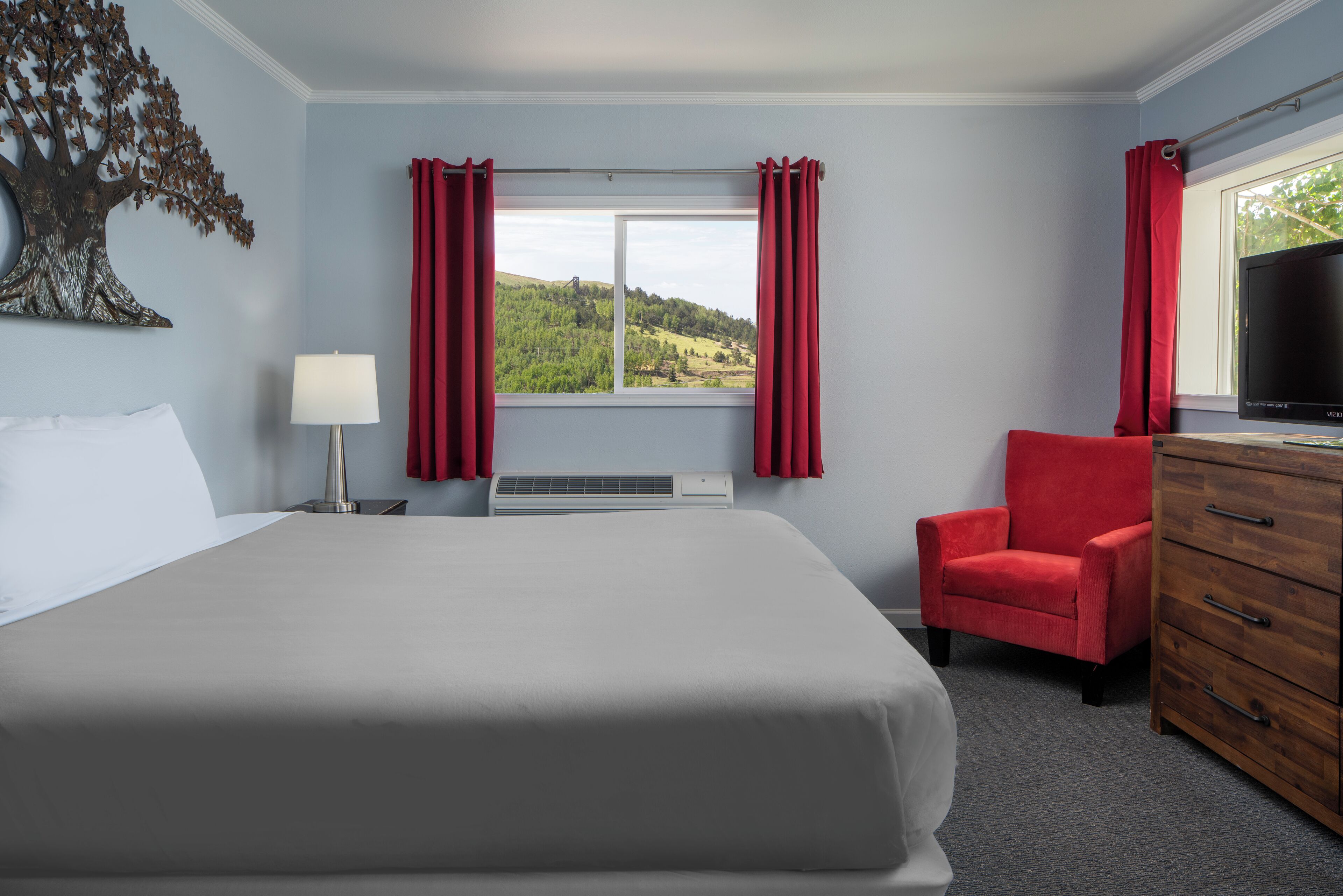 Ein Hotelzimmer mit einem großen Bett, einem roten Sessel, einer Holzkommode, einem Fernseher und einem Fenster mit roten Vorhängen.