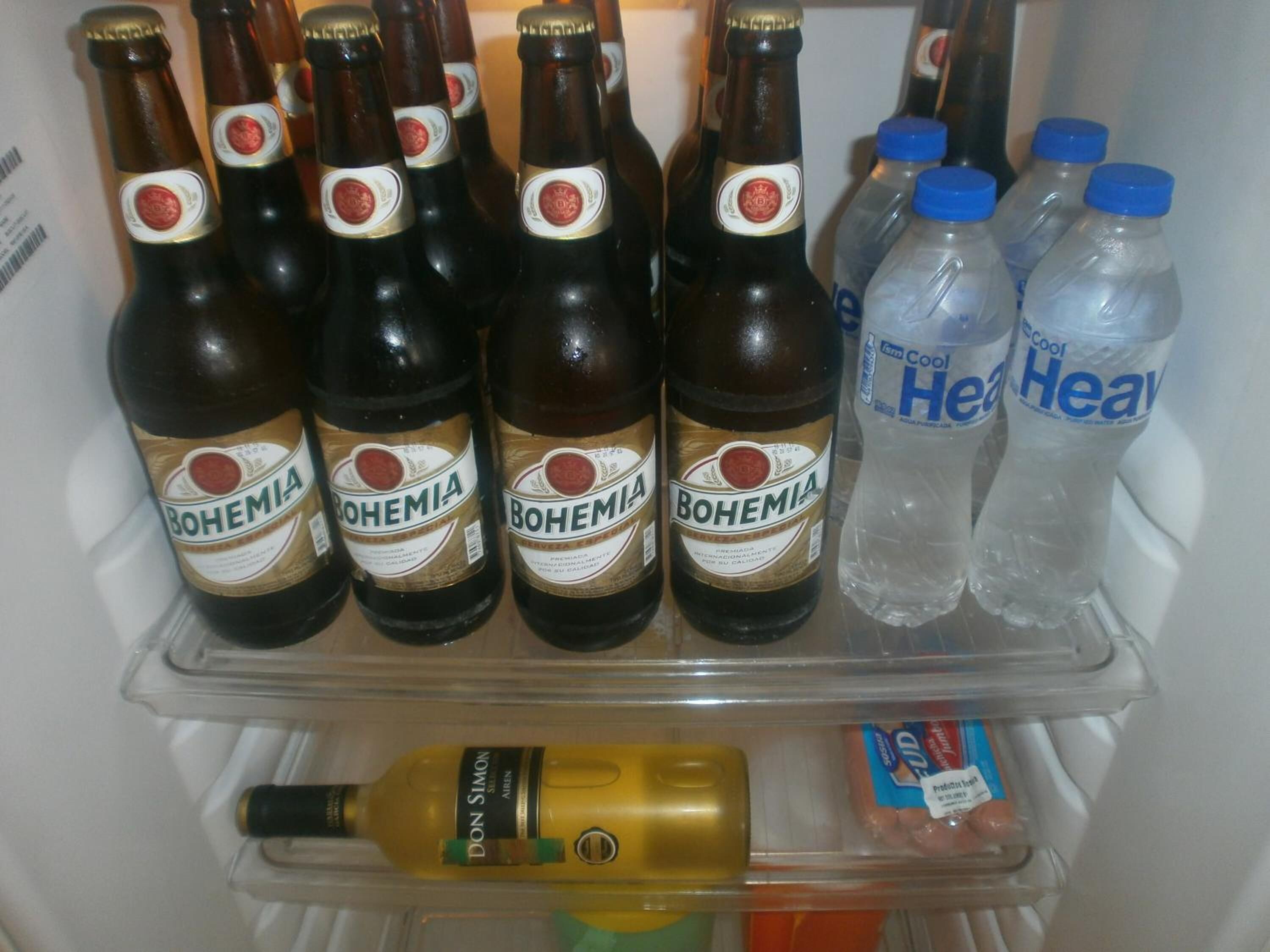 A refrigerator containing multiple bottles of Bohemia beer, two bottles of 'Cool Heavy' water, a bottle of Don Simon brand olive oil, and some packaged food items.