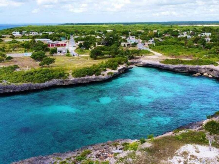 Uma área costeira com mar azul cristalino, uma pequena enseada e alguns edifícios dispersos.
