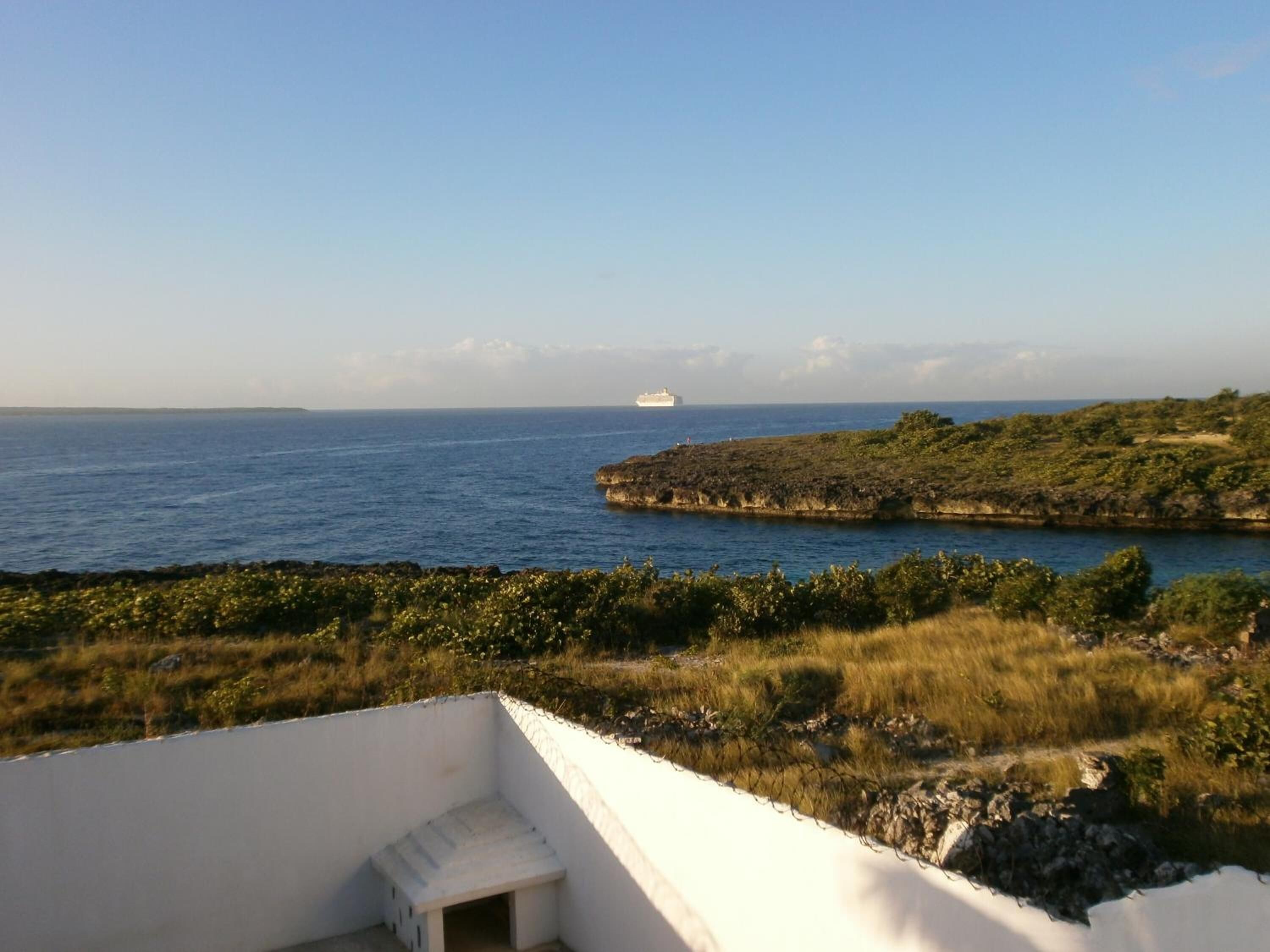 Un paisaje costero con un edificio blanco, un barco a lo lejos y un promontorio rocoso con vegetación.