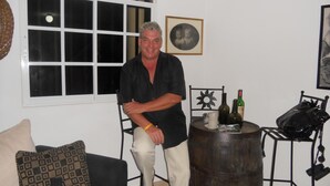 Um homem de camisa preta e calças bege sentado numa cadeira em um quarto com parede branca, uma janela com moldura branca, uma mesa redonda de madeira com garrafas e uma cadeira com desenho de sol.