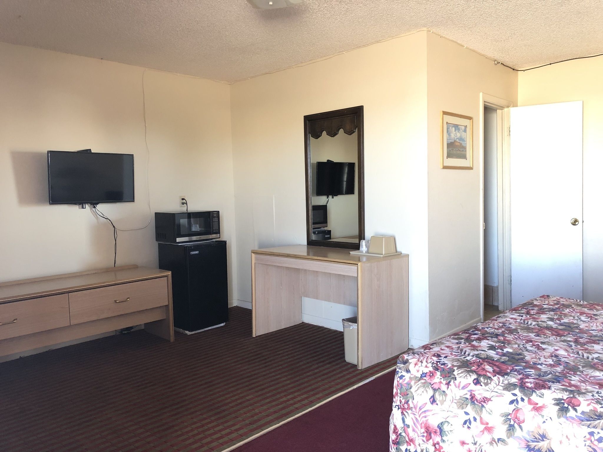 A hotel room with a bed, a desk with a mirror, a television mounted on the wall, and a small refrigerator.
