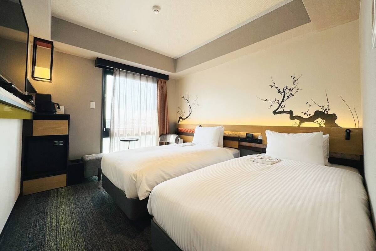 Ein Hotelzimmer mit zwei Betten, einem Schreibtisch, einem Stuhl und einem Fenster mit Vorhängen.