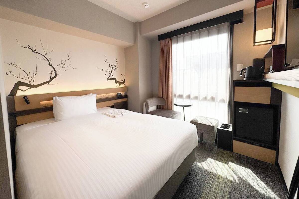 Ein Hotelzimmer mit einem großen Bett, einem Schreibtisch mit Stuhl, einem kleinen Tisch, einem Spiegel und einem Fenster mit Vorhängen.