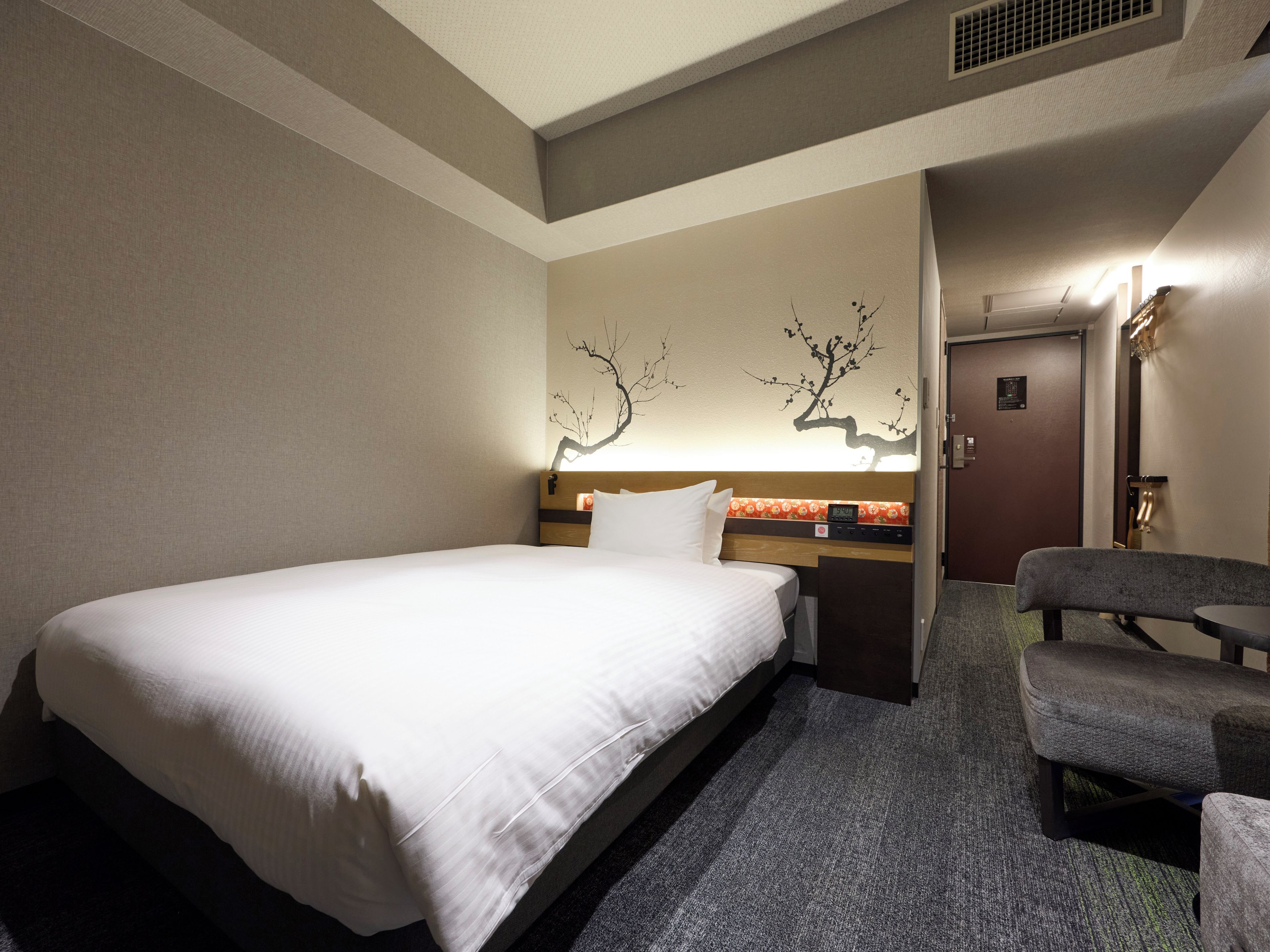 A hotel room with a bed, a chair, a nightstand, and a wall-mounted lamp.