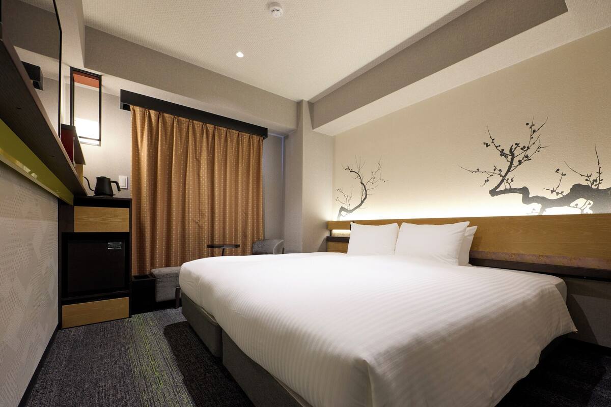 Ein Hotelzimmer mit einem großen Bett, einem Schreibtisch mit Computer, einem Sessel und einem Fenster mit Vorhängen.