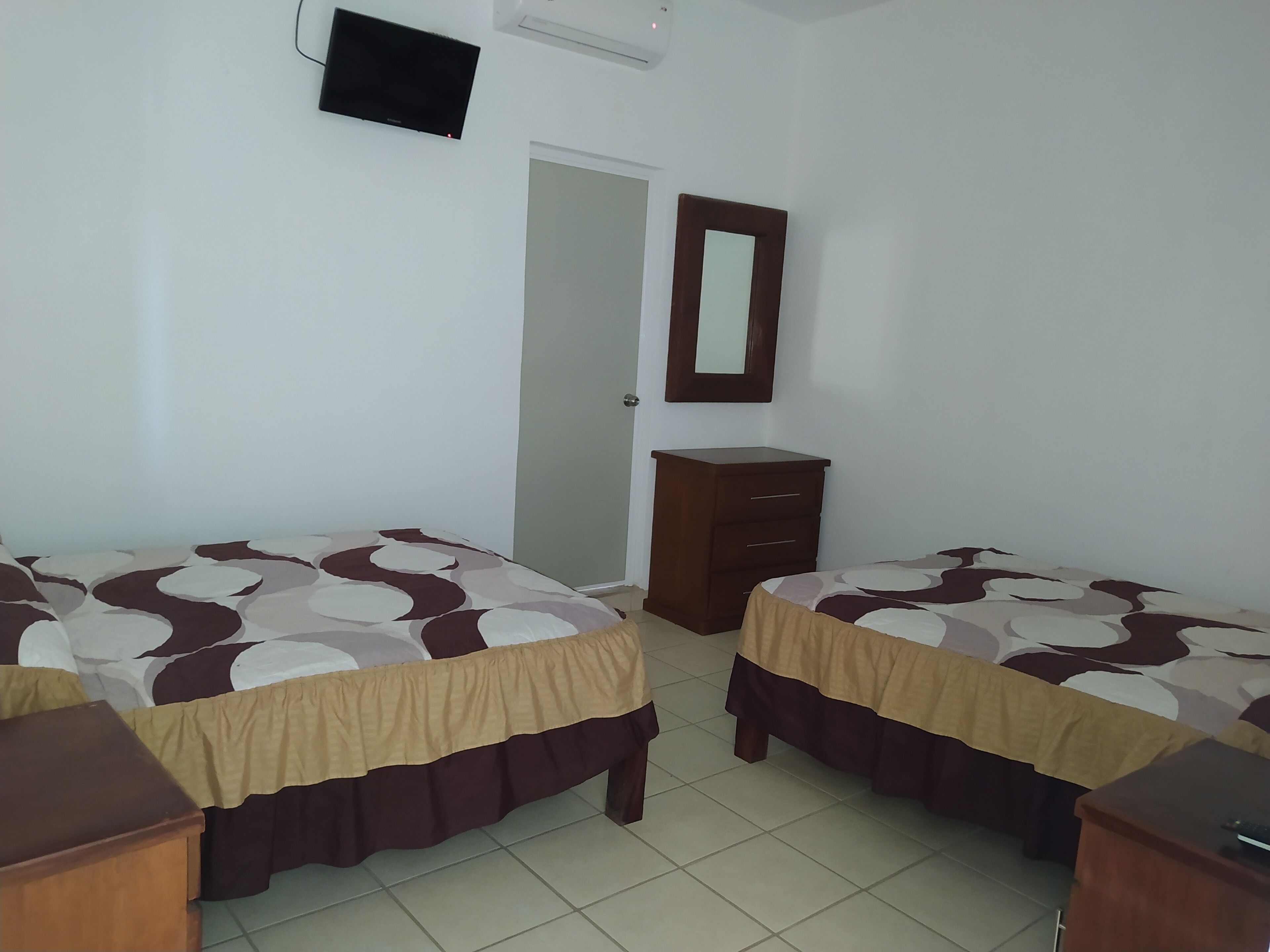 Une chambre avec deux lits, une commode en bois, un miroir et un téléviseur fixé au mur.