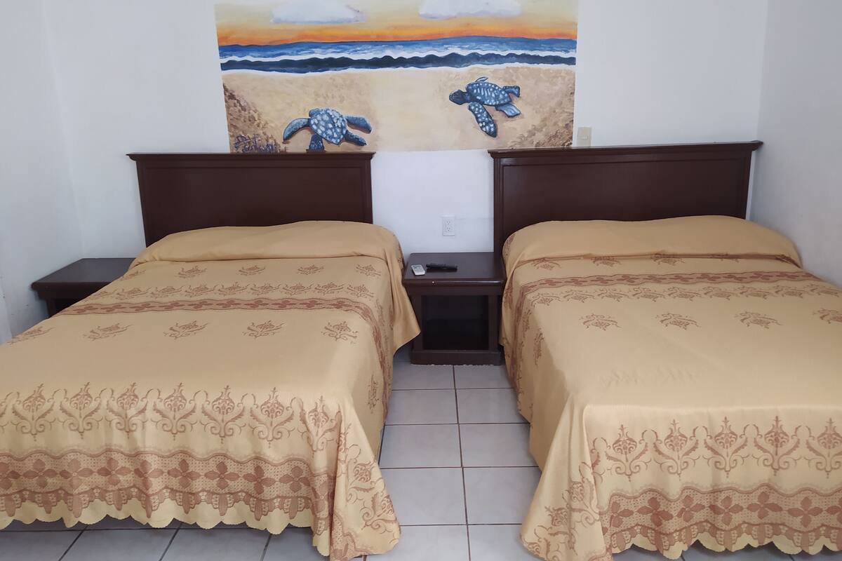 Ein Zimmer mit zwei Betten, jedes mit einem gemusterten Überwurf, einem Nachttisch mit Fernbedienung und einem Wandbild einer Schildkröte am Strand.