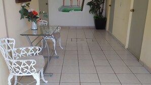 A hallway with a tiled floor, white metal chairs, a glass table, a painting of a gazebo, and a potted plant. - Hotel Lulu (Tondoroque)