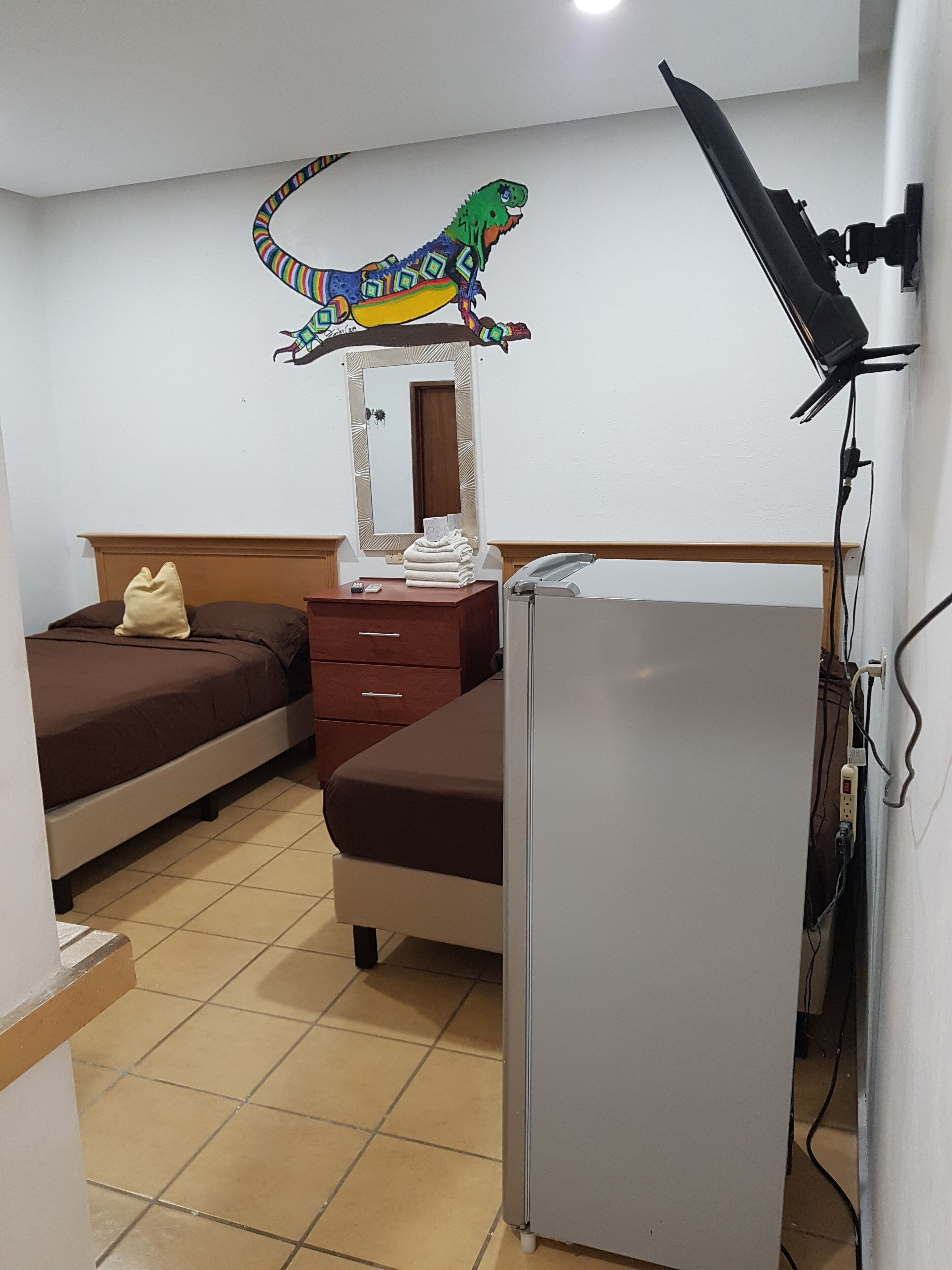 A hotel room with a bed, a desk, a mirror, a refrigerator, and a wall-mounted television.