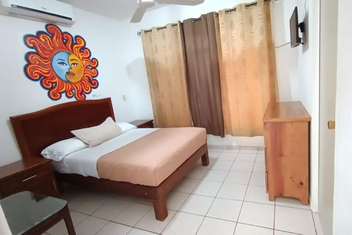 Ein Schlafzimmer mit einem Bett, einem Nachttisch aus Holz, einem kleinen Holztisch, einem an der Wand montierten Fernseher und einem Deckenventilator.