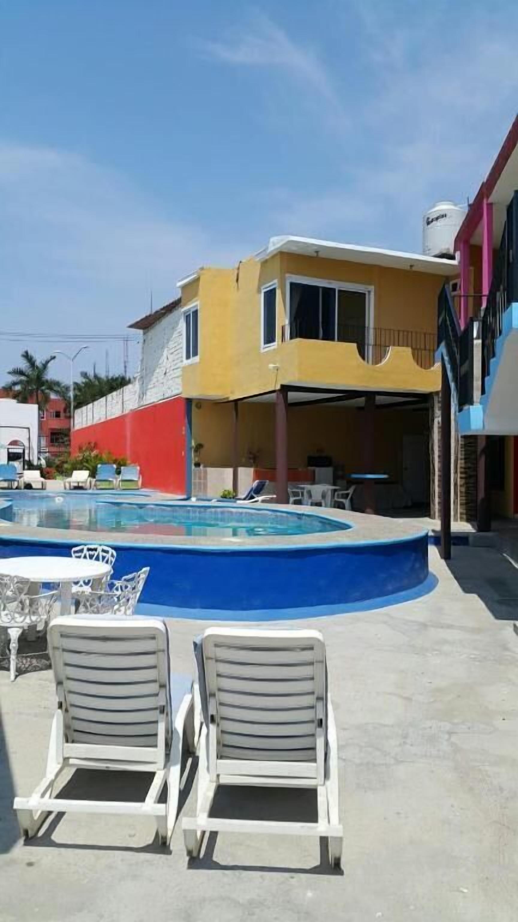 Zona de piscina con dos sillones de descanso, un edificio amarillo y un muro rojo, y un cielo azul.