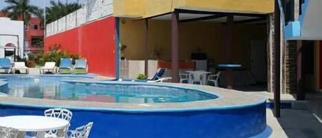 Zona de piscina con dos sillones de descanso, un edificio amarillo y un muro rojo, y un cielo azul.
