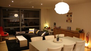 A modern living room with a sofa, dining table, and chairs. There is a large window, a red chair, and a plant on the table. - Casa Nina (Ostend)