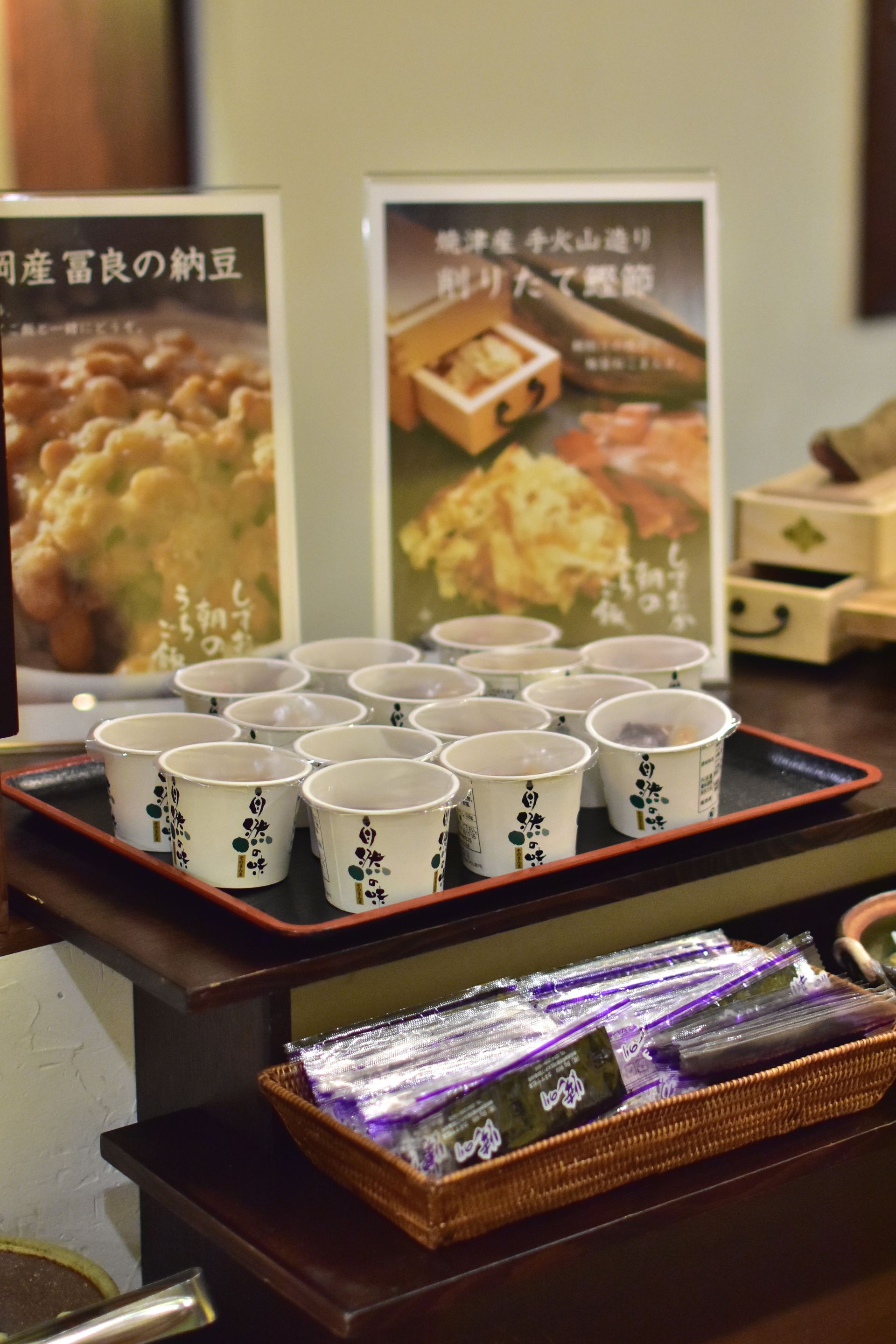 A display of disposable cups with Japanese characters, a wicker basket with packaged items, and two posters with Japanese text.