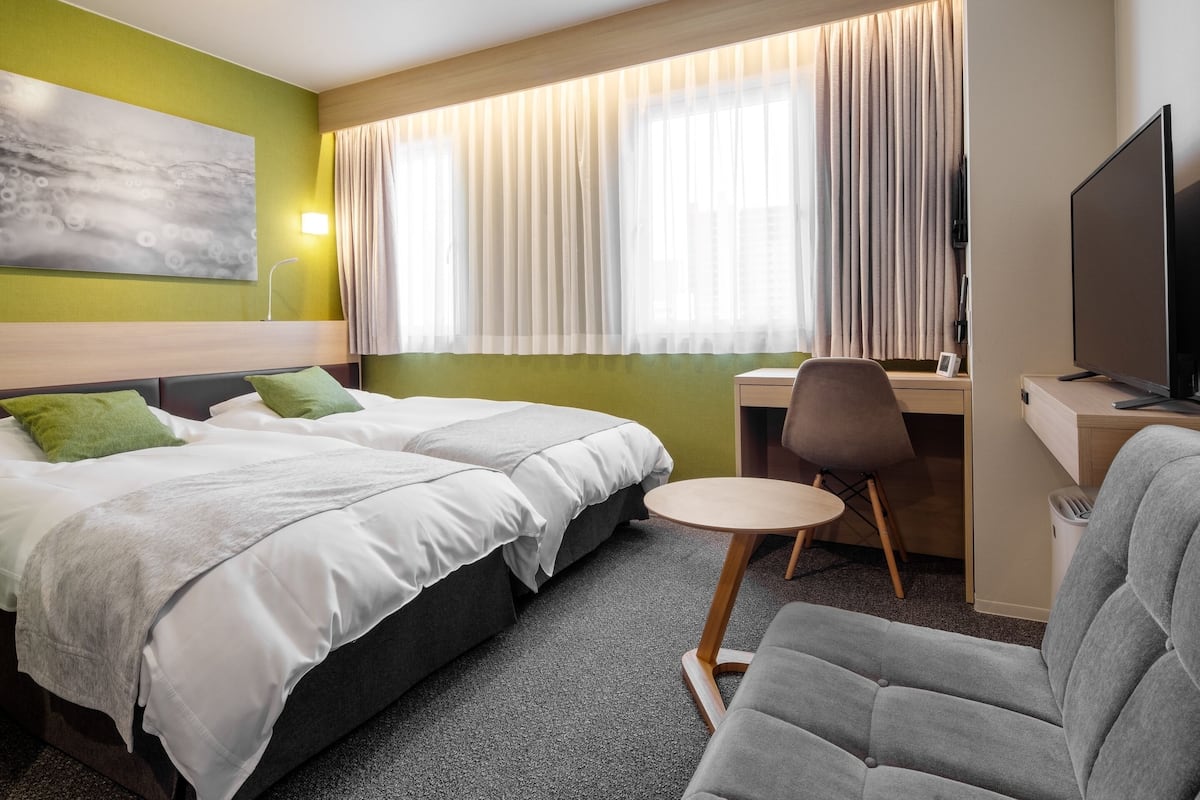 A hotel room with two beds, a desk, a chair, a television, and a large window with curtains.