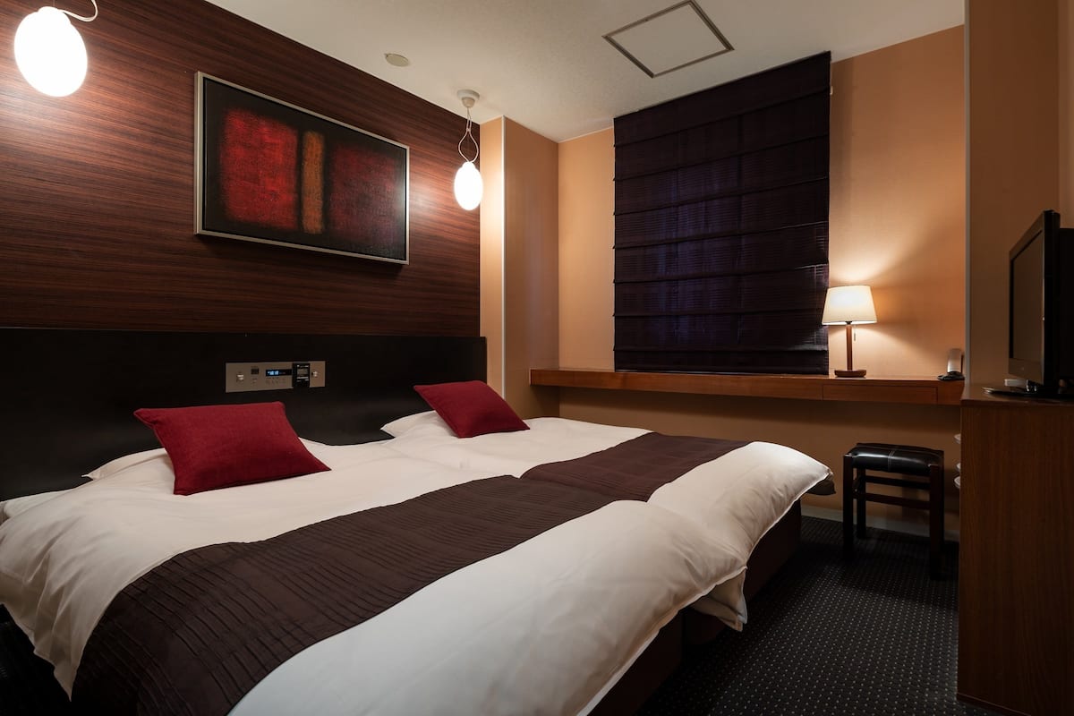 A hotel room with a large bed, two red pillows, a headboard, a bedside table with a lamp, a small stool, and a television on a stand.