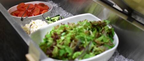 A stainless steel buffet setup with various food items including a bowl of sliced tomatoes, a bowl of green salad, and a container of rice.