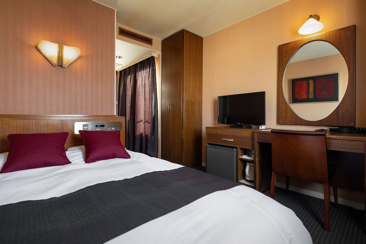 A hotel room with a bed, a desk, a television, a mirror, and a wall-mounted light fixture.