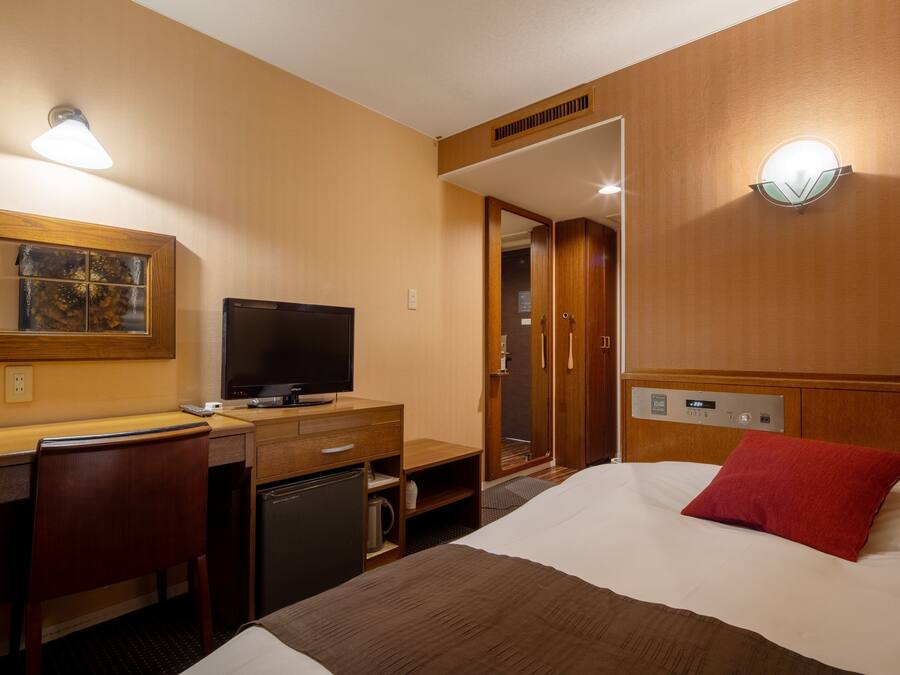 A hotel room with a bed, a desk with a television, a small refrigerator, and a shower area.